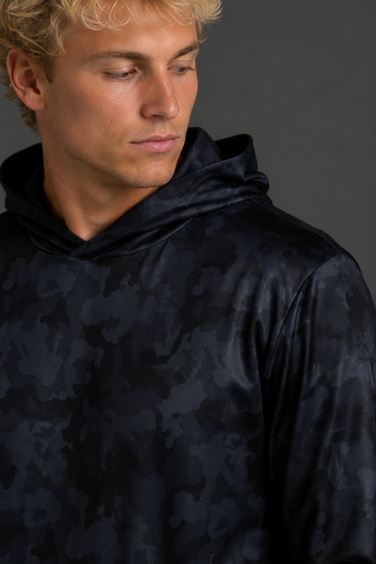 A blonde man wearing a dark navy hooded sweatshirt with a subtle tonal abstract pattern, standing against a solid dark gray studio background. || Black Camo