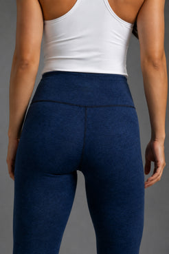 Rear view of a person wearing high-waisted heathered navy blue leggings and a white sports bra against a neutral gray background. || Navy