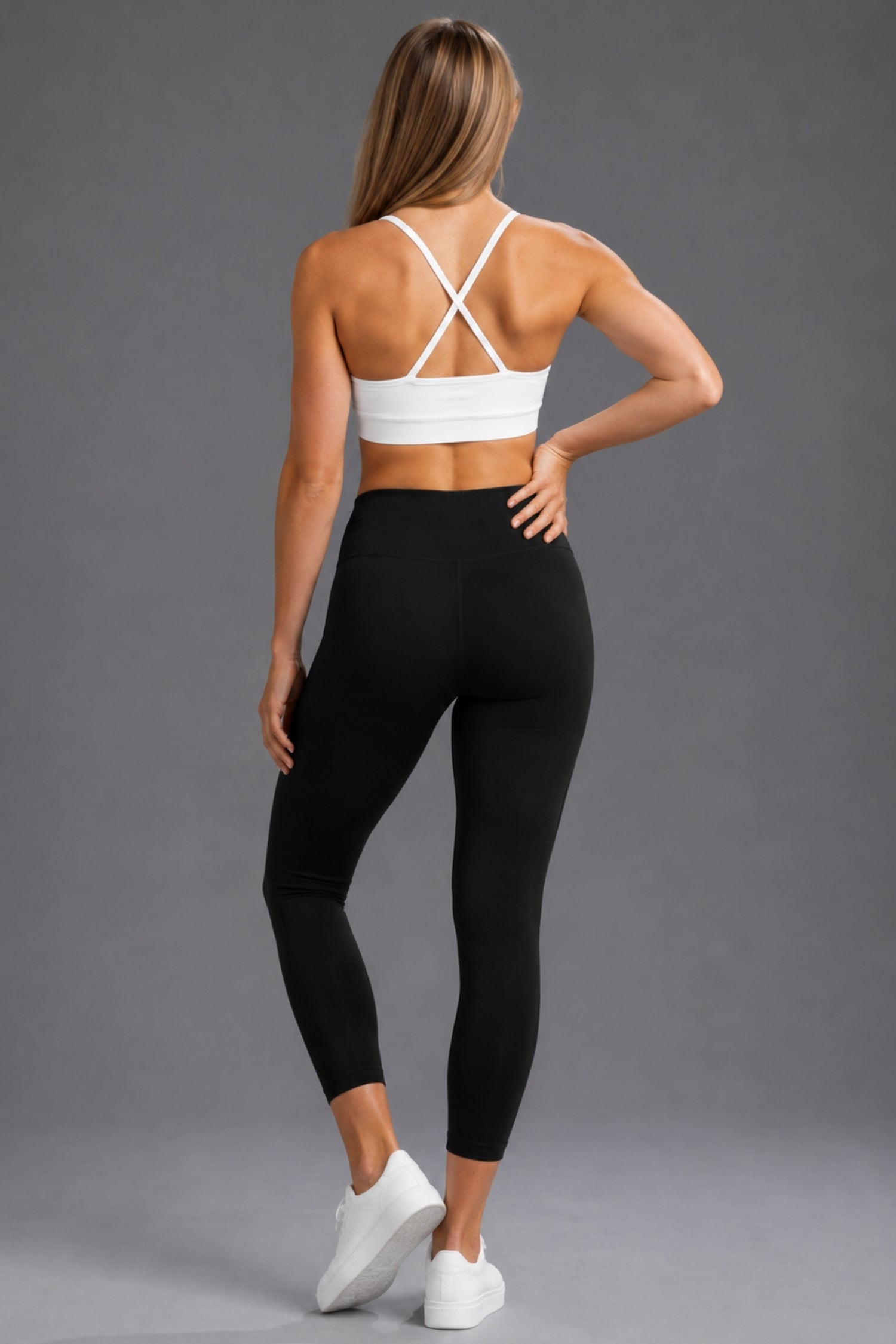 A woman shown from the back wearing a white sports bra with crisscross straps and black high-waisted leggings, standing against a solid gray studio background. || Black Heather