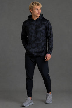A smiling blonde man wearing a dark navy hooded sweatshirt with a subtle tonal abstract pattern, standing against a solid dark gray studio background. || Black Camo