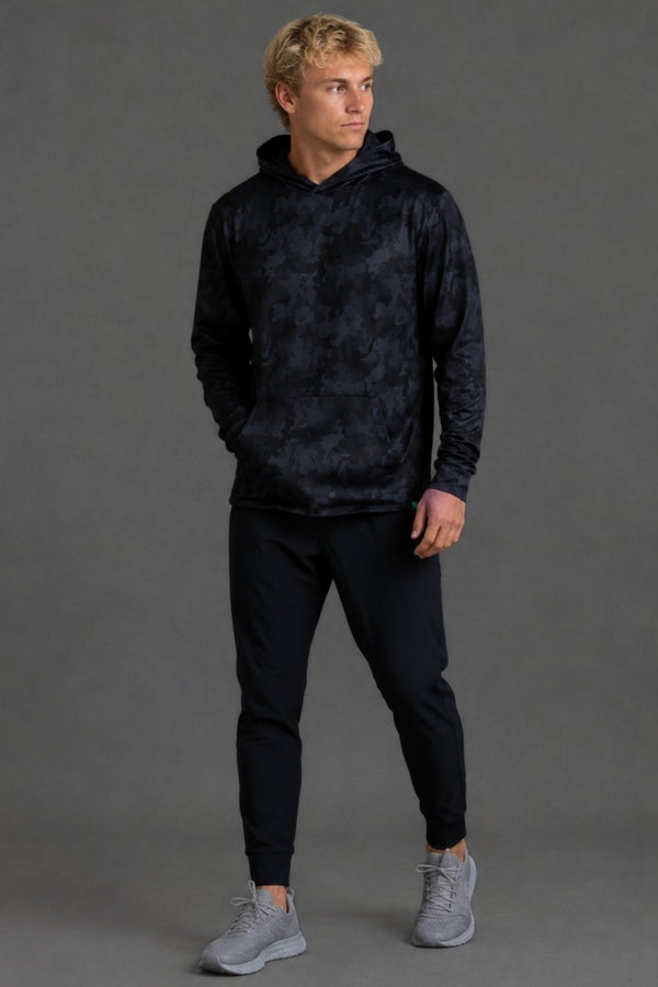 A smiling blonde man wearing a dark navy hooded sweatshirt with a subtle tonal abstract pattern, standing against a solid dark gray studio background. || Black Camo