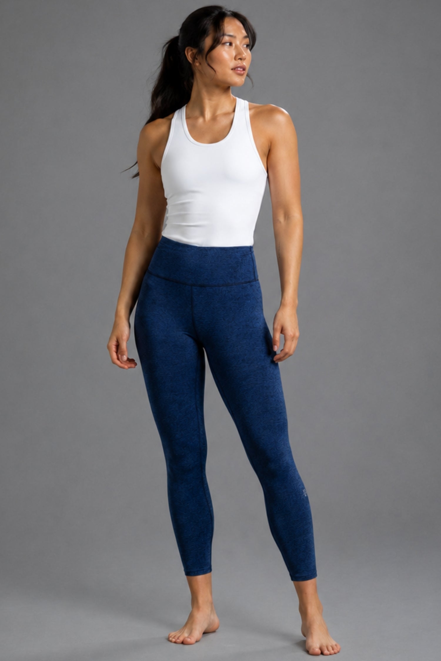 A woman modeling high-waisted, ankle-length leggings in a deep navy blue color, paired with a white tank top, against a solid gray studio background.