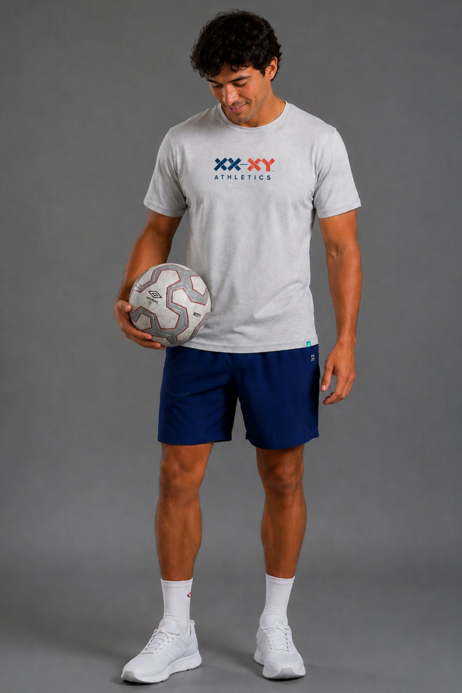 A man modeling a light gray short-sleeve athletic t-shirt with a blue and red 'XX-XY ATHLETICS' logo, paired with navy blue athletic shorts, white socks, and white sneakers, holding a soccer ball against a solid gray studio background. || Light Heather Gray - 1