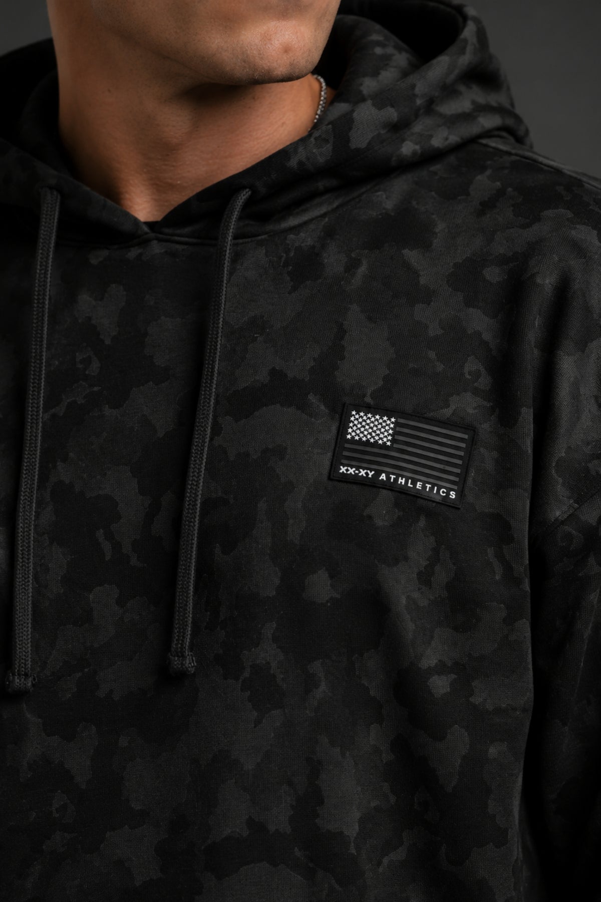 Close-up of a black camo hoodie featuring a monochrome American flag XX-XY Athletics patch on the chest.