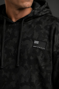 Close-up of a black camo hoodie featuring a monochrome American flag XX-XY Athletics patch on the chest. || Black Camo