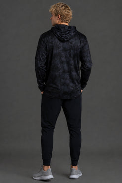 A man viewed from behind wearing a dark navy hooded sweatshirt with a subtle tonal pattern, black jogger pants, and gray sneakers, standing against a solid dark gray studio background. || Black Camo