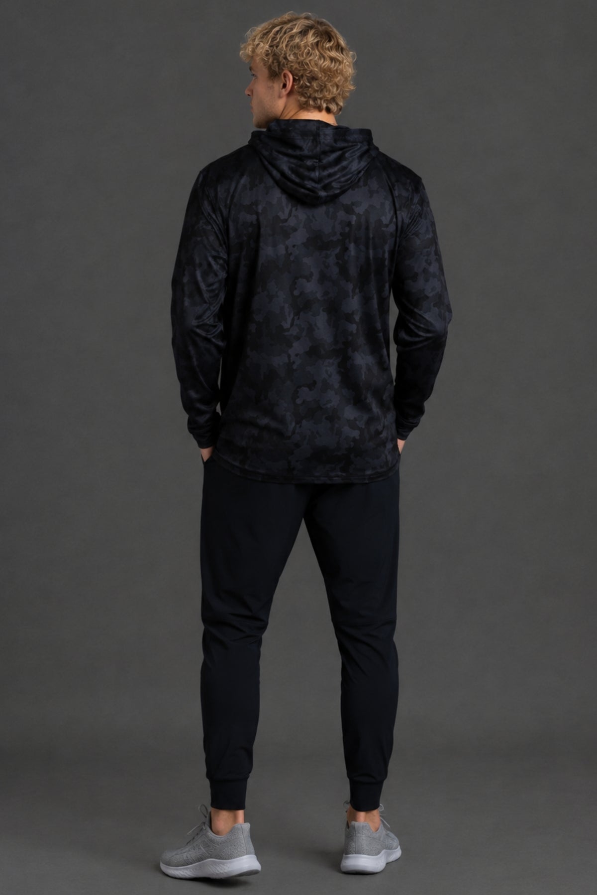 A man viewed from behind wearing a dark navy hooded sweatshirt with a subtle tonal pattern, black jogger pants, and gray sneakers, standing against a solid dark gray studio background. || Black Camo