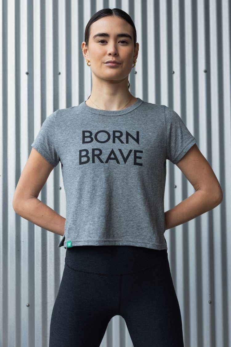Person wearing a gray 'Born Brave' t-shirt against a corrugated metal background || heather gray