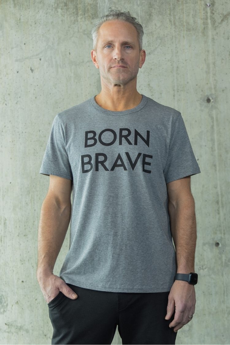 Man wearing a gray t-shirt with 'BORN BRAVE' text against a concrete wall.