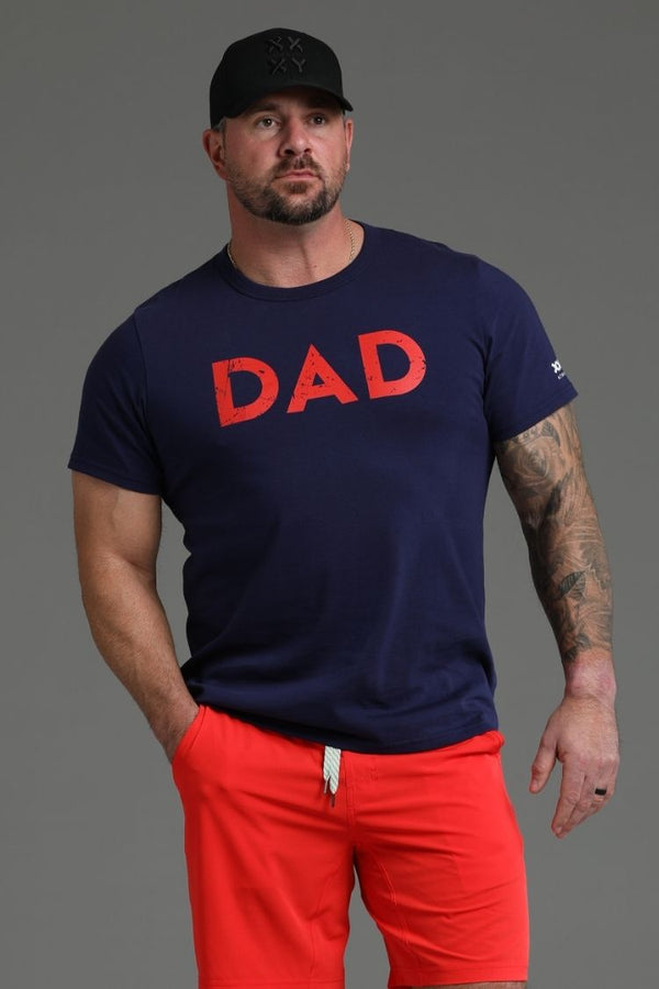 Man wearing a navy t-shirt with 'DAD' in red, red shorts, and a black cap on a gray background || dark navy