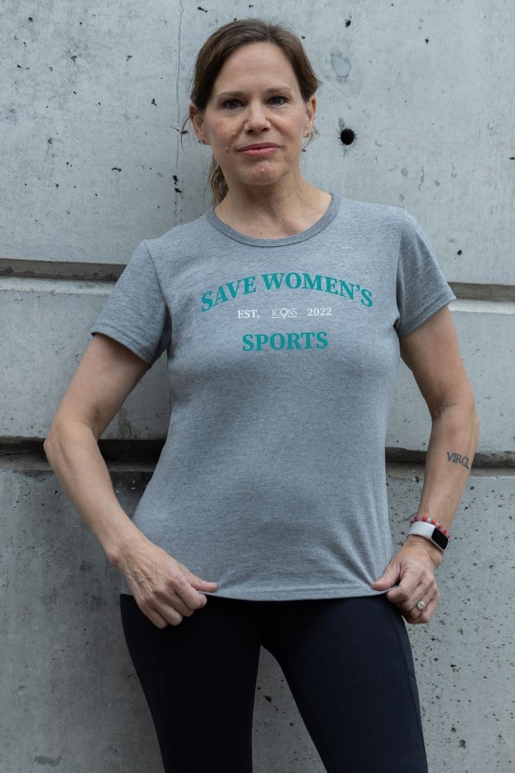 Woman wearing a gray t-shirt with 'Save Women's Sports' text against a concrete wall.