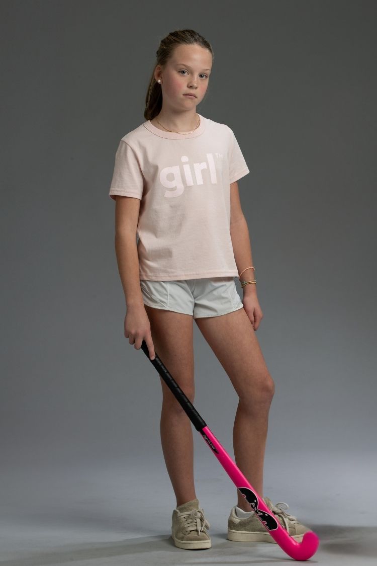 Young girl holding a pink and black hockey stick, wearing a 'girl' t-shirt on a gray background || light pink