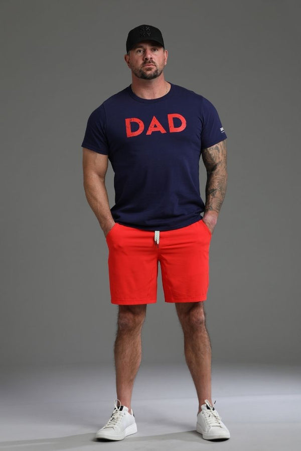 Man wearing a navy t-shirt with 'DAD' in red, red shorts, and white sneakers on a gray background || dark navy