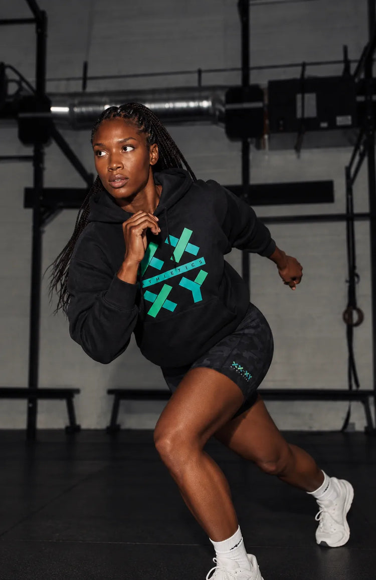 Female athlete wearing a black XX-XY Athletics hoodie with a teal logo, black athletic shorts, and white sneakers poses in a dynamic running stance inside a gym.