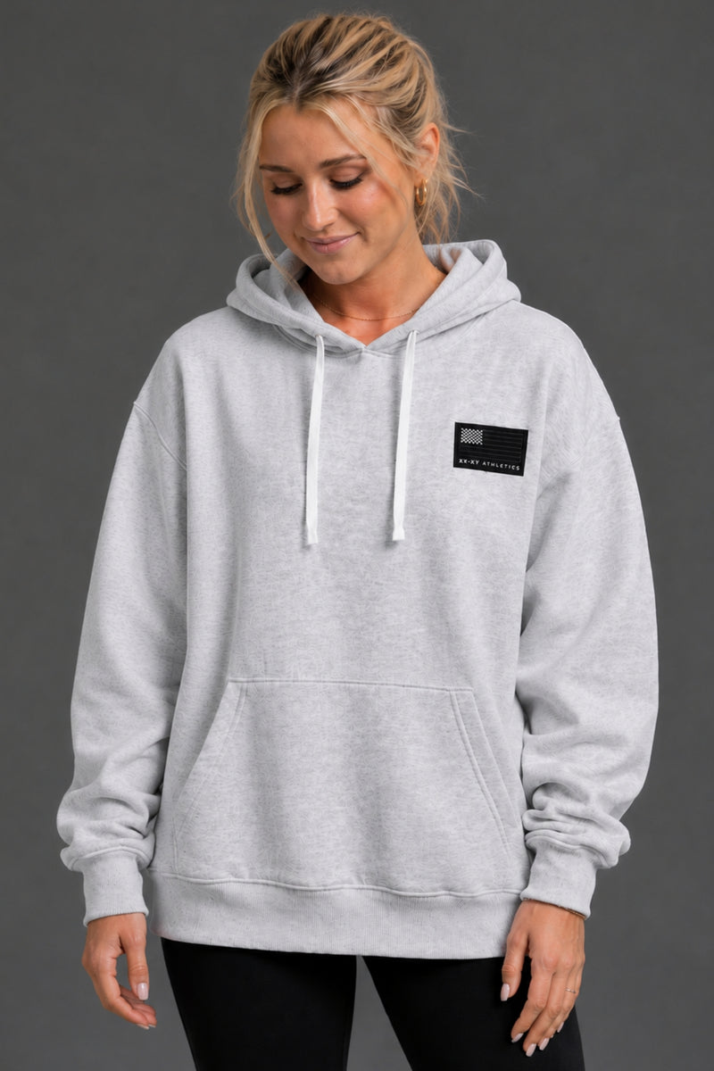 Woman wearing a gray hoodie with a small patch on a dark background || Light Heather Gray