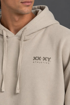 Male model wearing a beige XX-XY Athletics logo hoodie with mocha shorts against a gray background. || Taupe