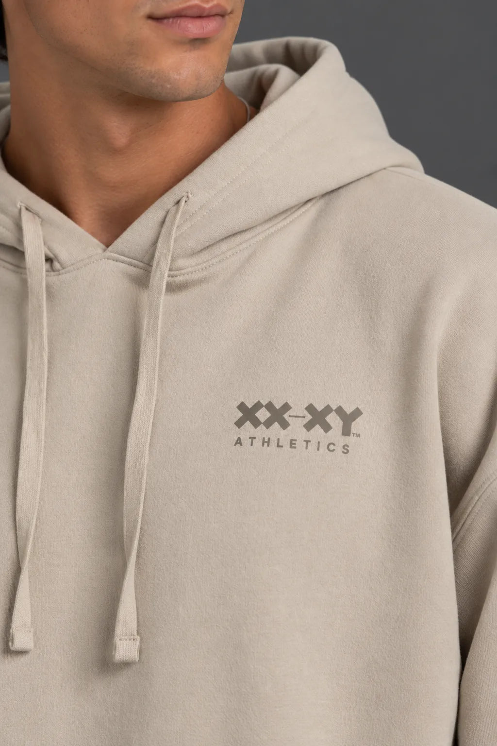 Male model wearing a beige XX-XY Athletics logo hoodie with mocha shorts against a gray background. || Taupe