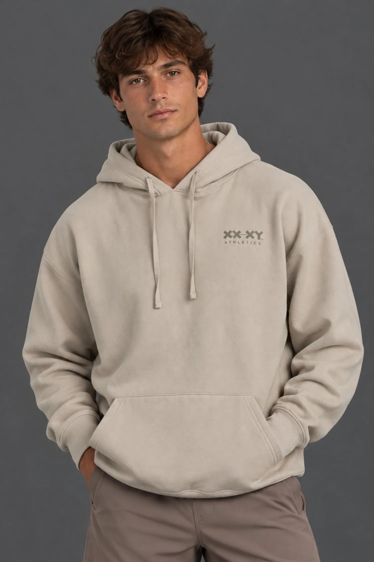 Male model wearing a beige XX-XY Athletics logo hoodie with mocha shorts against a gray background. || Taupe