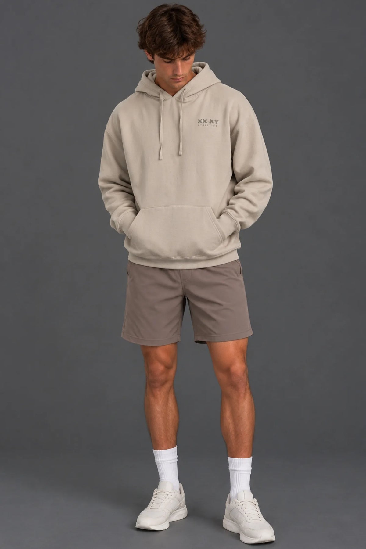 Male model wearing a beige XX-XY Athletics hoodie with mocha athletic shorts against a gray studio background.