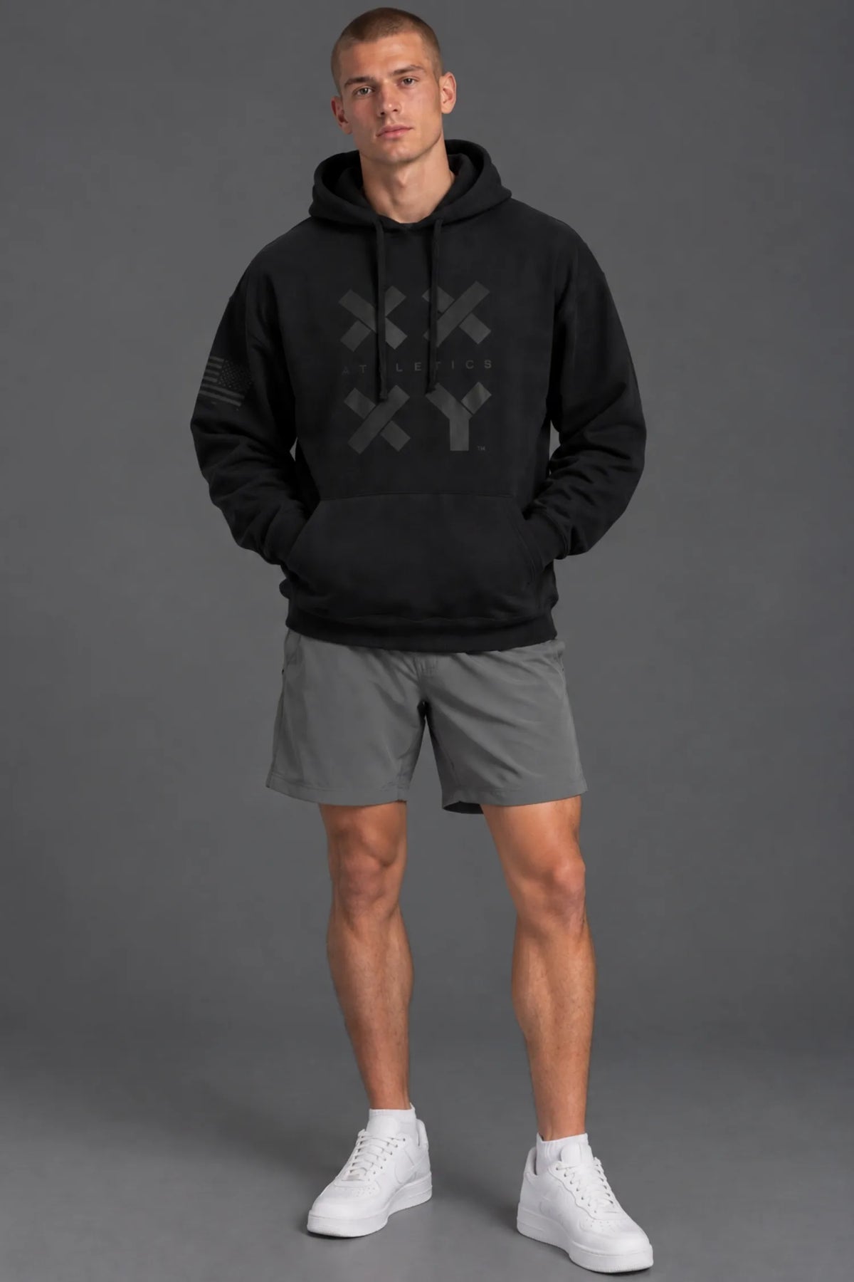 Male model wearing a black XX-XY Athletics graphic hoodie with gray athletic shorts against a gray studio background. || Black