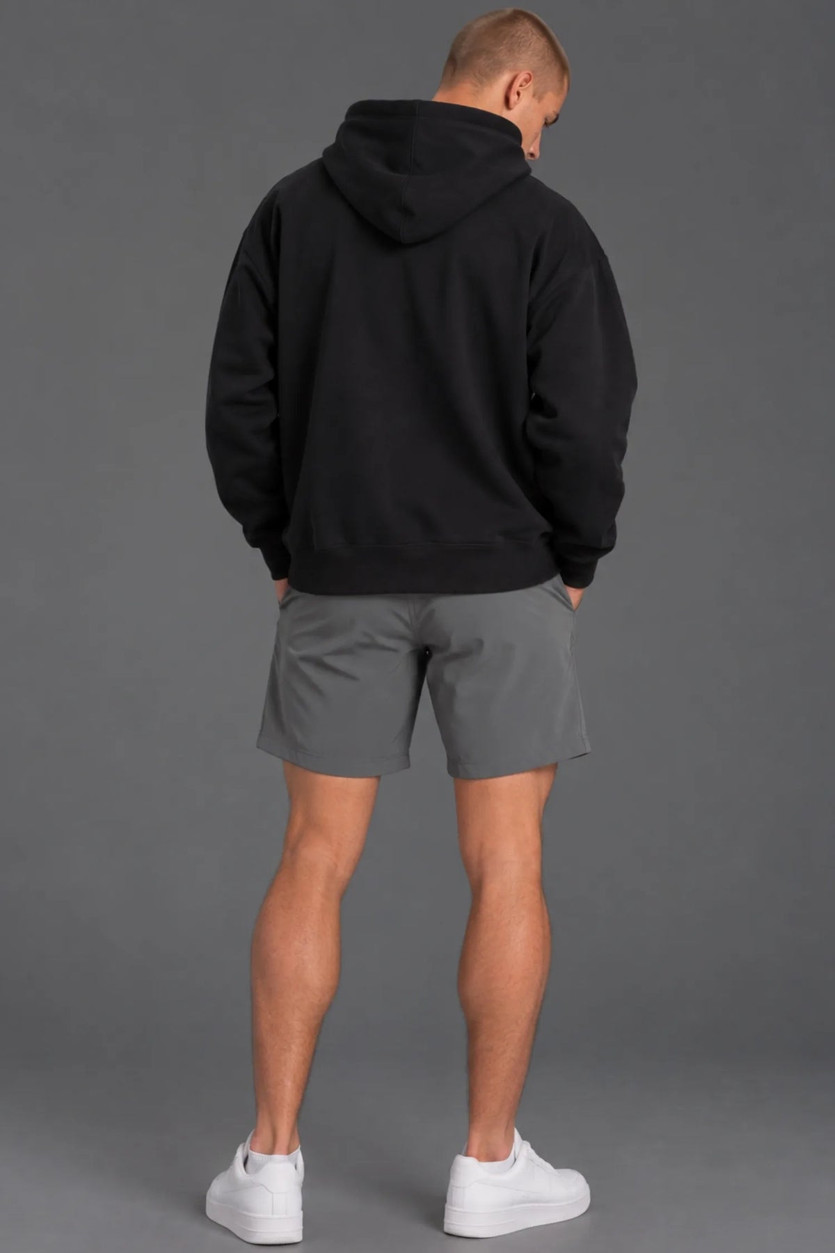 Back view of a male model wearing a blank black hoodie with gray athletic shorts. || Black