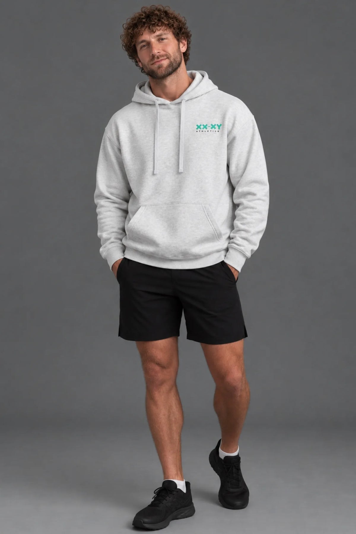 Male model wearing a heather gray XX-XY Athletics hoodie, black shorts, ankle socks, and black sneakers.