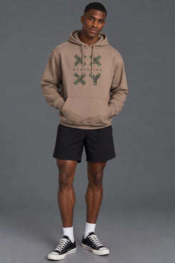 Male model wearing a mocha XX-XY Athletics camo graphic hoodie with black athletic shorts against a gray studio background.|| Mocha