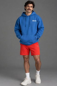 Male model wearing a blue XX-XY Athletics hoodie with red athletic shorts against a gray studio background. || Cobalt Blue
