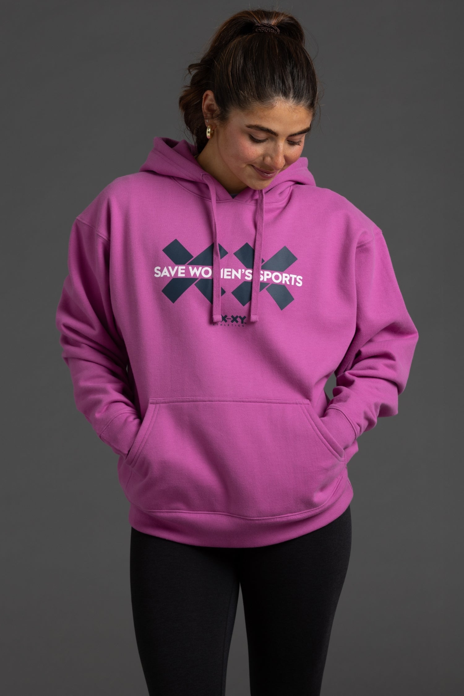 A woman wearing a bright pink pullover hoodie with a dark navy graphic on the chest that reads 'SAVE WOMEN'S SPORTS' and 'SKYX', paired with black leggings, against a solid dark gray studio background.