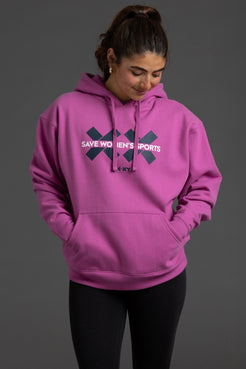 A woman wearing a bright pink pullover hoodie with a dark navy graphic on the chest that reads 'SAVE WOMEN'S SPORTS' and 'SKYX', paired with black leggings, against a solid dark gray studio background. || Bright Fuchsia