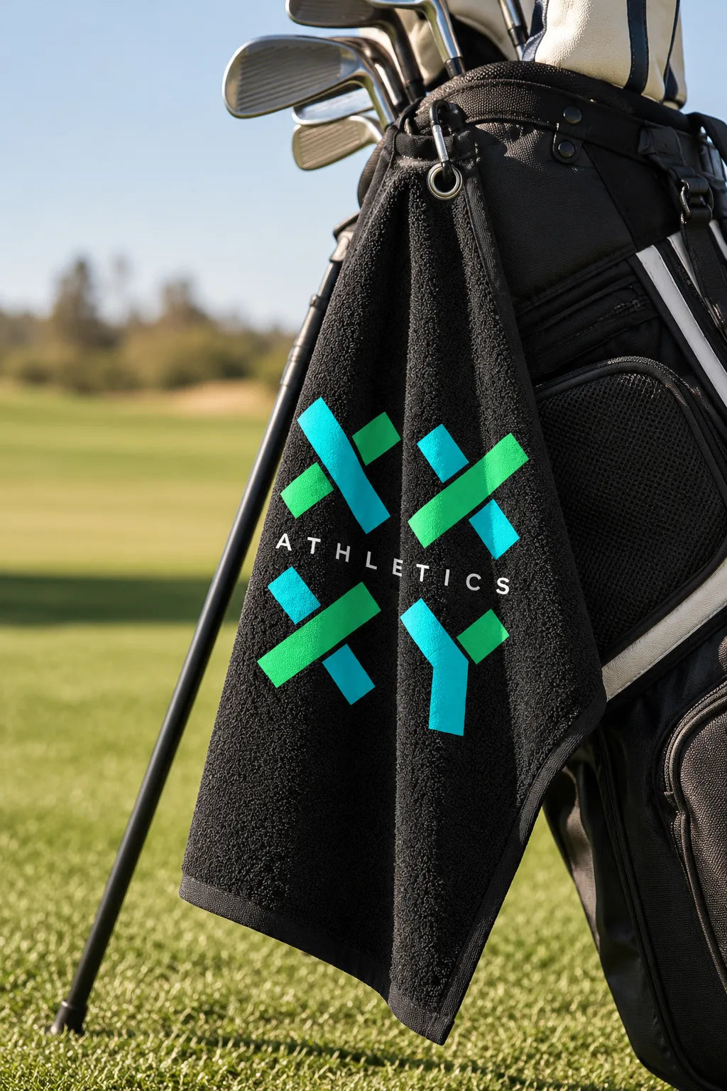 Black XX-XY Athletics golf towel hanging from a golf bag on the fairway, featuring the turquoise and green logo with golf clubs in the background