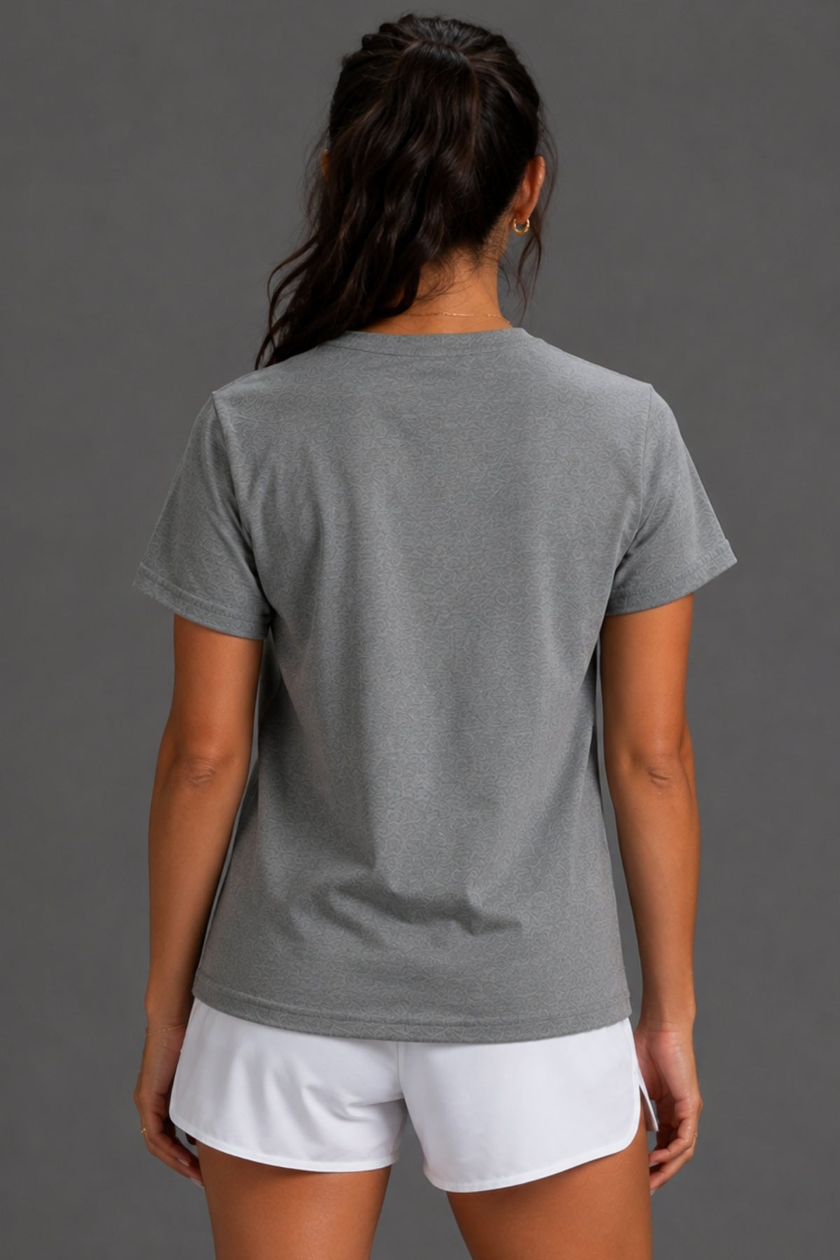A woman viewed from the back wearing a heather gray short-sleeve t-shirt and white athletic shorts, set against a solid dark gray studio background.