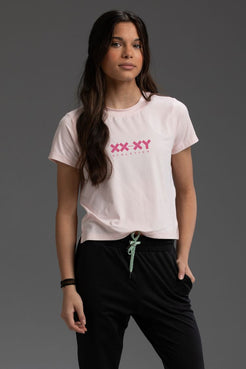 Woman wearing a light pink t-shirt with 'XXXY' logo on a gray background || Light Pink Heather