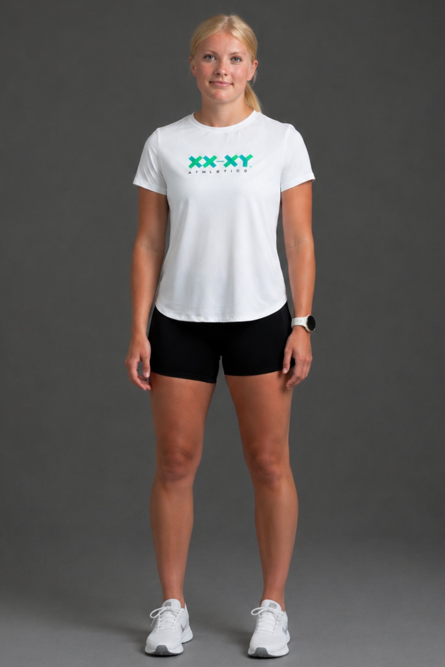 A woman modeling a white short-sleeve athletic t-shirt with a green 'XX XY ATHLETICS' logo on the chest, paired with black athletic shorts and white sneakers, against a solid gray studio background. || White