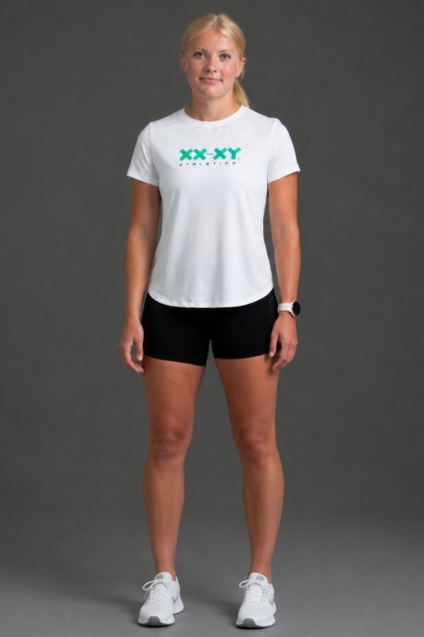 A woman modeling a white short-sleeve athletic t-shirt with a green 'XX XY ATHLETICS' logo on the chest, paired with black athletic shorts and white sneakers, against a solid gray studio background. || White