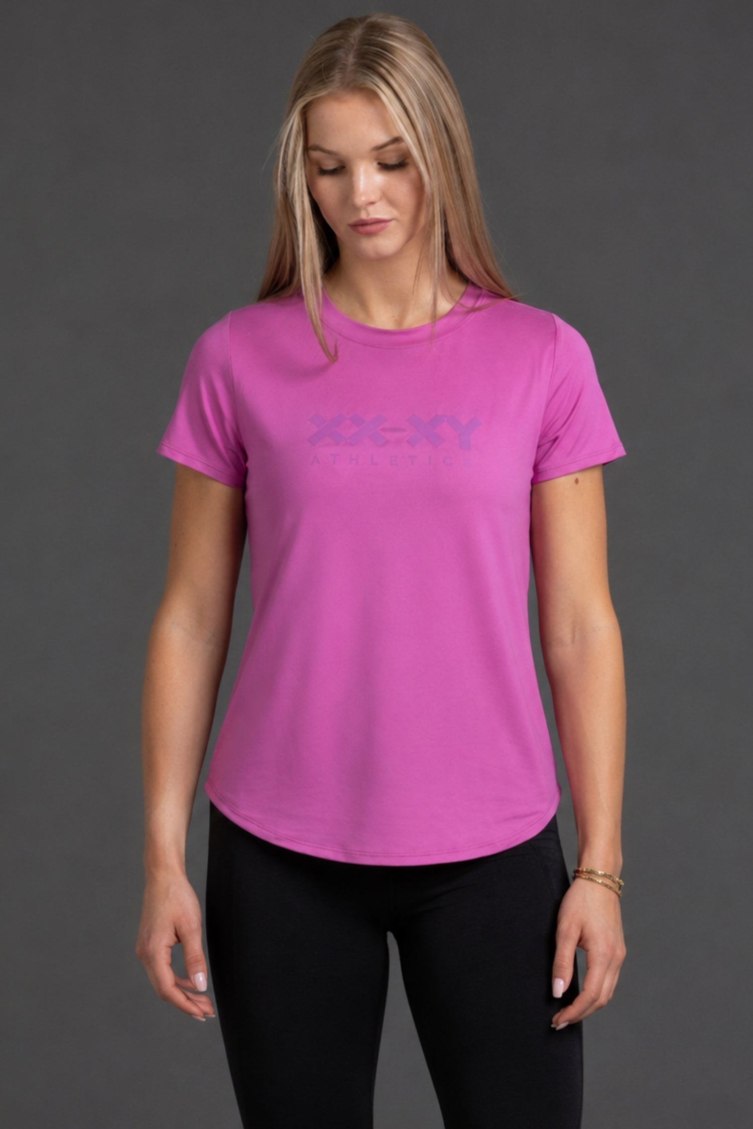 A woman wearing a bright pink short-sleeve athletic t-shirt with a subtle tonal logo across the chest, paired with black leggings, against a dark gray studio background.
