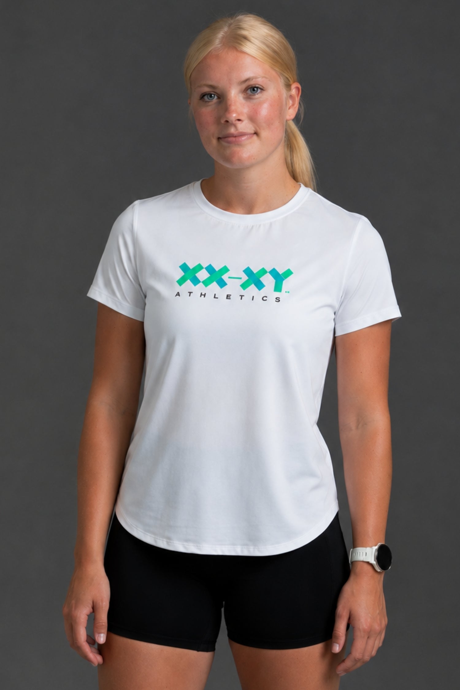 A woman modeling a white short-sleeve athletic T-shirt with a teal and black 'XX-XY ATHLETICS' logo on the chest, paired with black athletic shorts against a dark gray studio background.