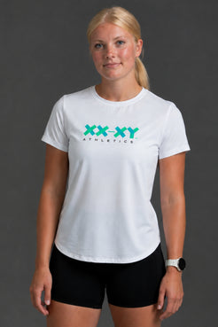 A woman modeling a white short-sleeve athletic T-shirt with a teal and black 'XX-XY ATHLETICS' logo on the chest, paired with black athletic shorts against a dark gray studio background. || White