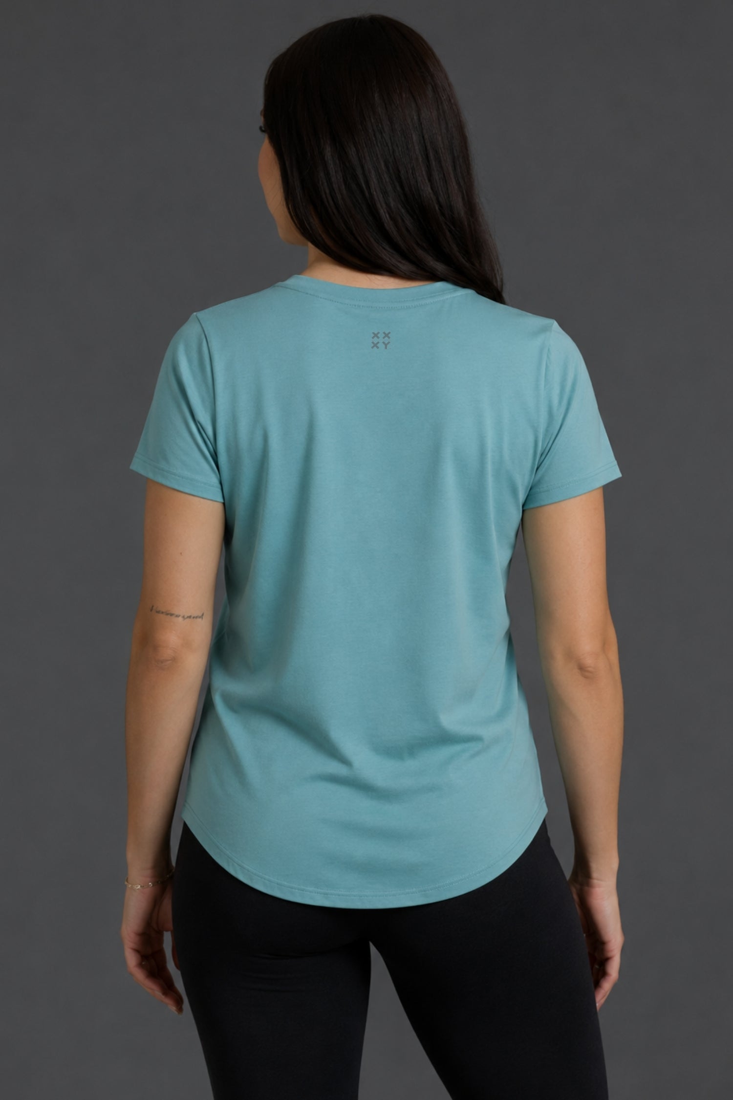 A woman viewed from the back wearing a light blue athletic short-sleeve t-shirt and black leggings against a dark gray studio background.