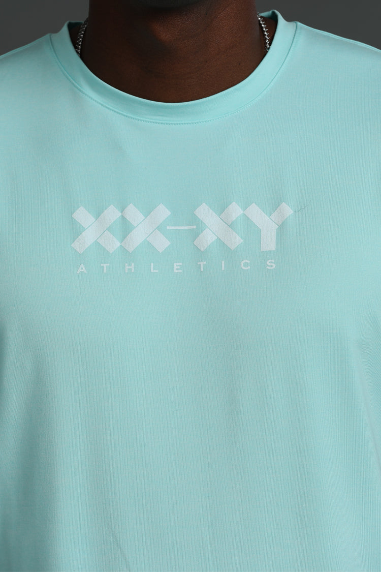 A close-up of a person wearing a light mint green crewneck t-shirt with a white 'XX-XY ATHLETICS' logo printed across the chest.