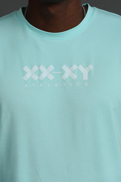A close-up of a person wearing a light mint green crewneck t-shirt with a white 'XX-XY ATHLETICS' logo printed across the chest. || Aqua Heather