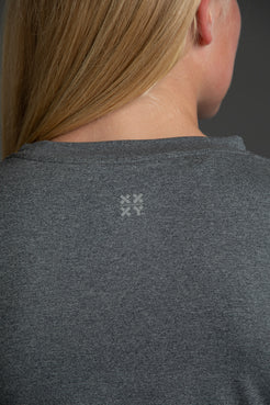 A woman modeling a heather gray athletic t-shirt with a white 'XX XY ATHLETICS' logo on the back against a dark gray studio background. || Dark Heather Gray