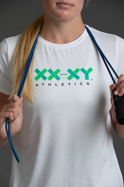 A woman wearing a white athletic t-shirt with a green and black 'XX-XY ATHLETICS' logo on the chest, holding blue resistance bands against a dark gray background. || White