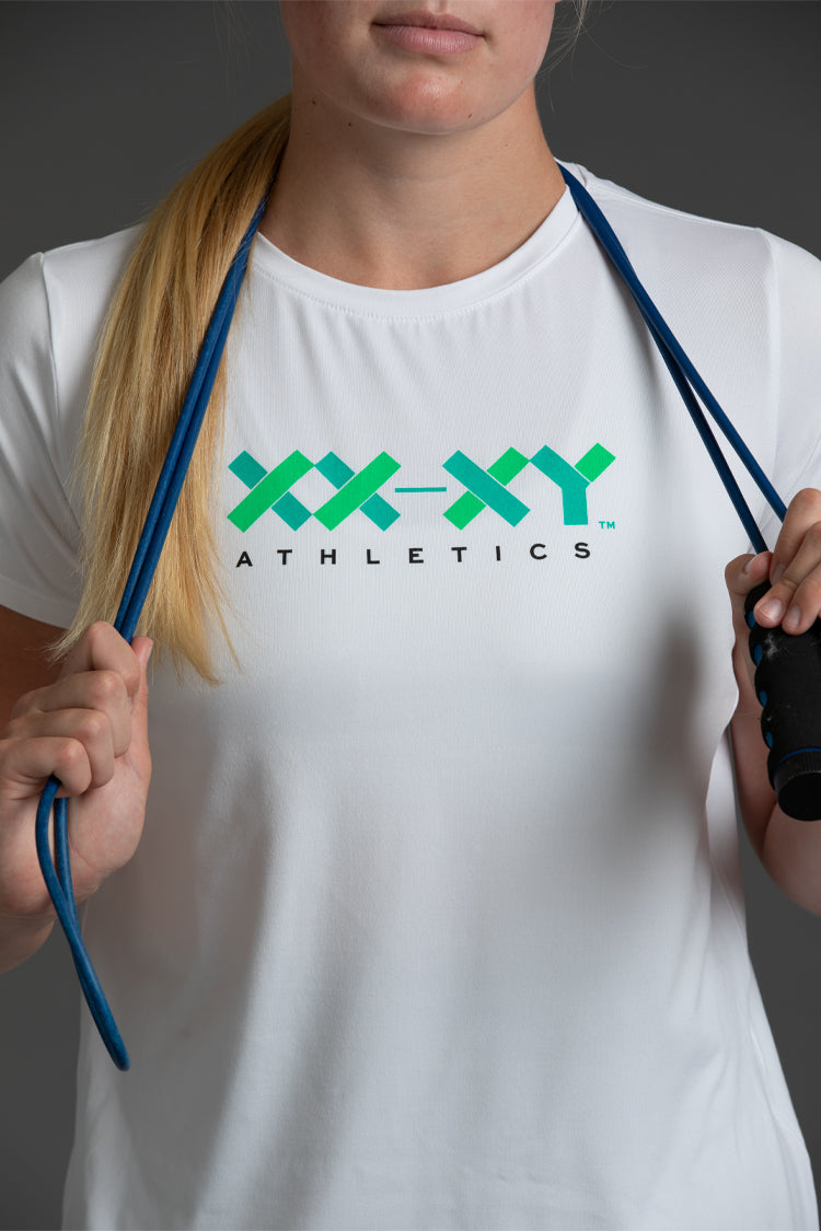A woman wearing a white athletic t-shirt with a green and black 'XX-XY ATHLETICS' logo on the chest, holding blue resistance bands against a dark gray background. || White