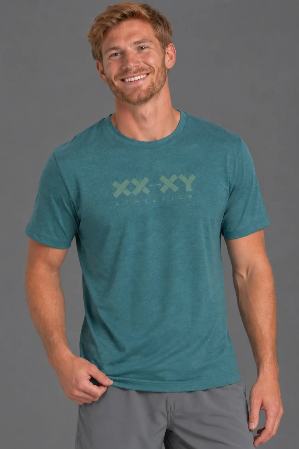 A smiling man wearing a teal heathered short-sleeve t-shirt with a light green 'XX-XY ATHLETICS' logo on the chest, paired with gray athletic shorts, against a solid gray studio background. || Teal Green