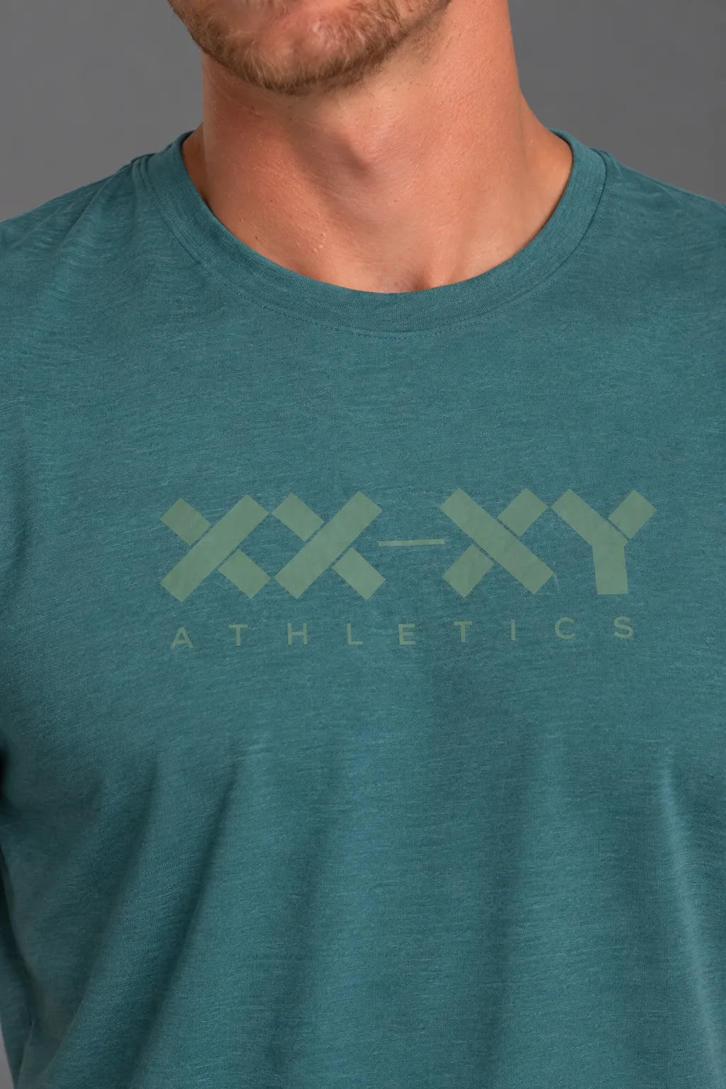 A close-up of a man wearing a heathered teal athletic t-shirt with a light sage green 'XX-XY ATHLETICS' logo printed across the chest.