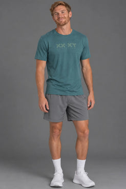 A man modeling a teal athletic t-shirt with a subtle 'XX XY' logo on the chest, paired with gray athletic shorts, white socks, and white sneakers, against a solid gray studio background. || Teal Green