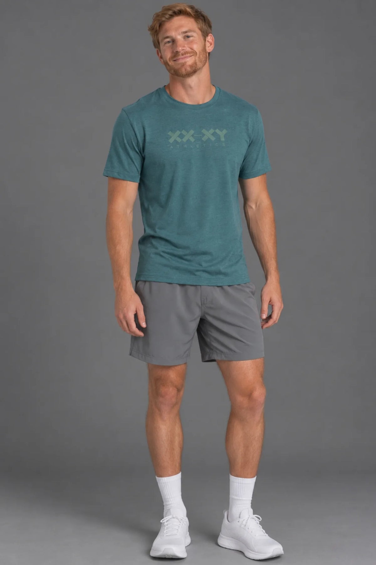 A man modeling a teal athletic t-shirt with a subtle 'XX XY' logo on the chest, paired with gray athletic shorts, white socks, and white sneakers, against a solid gray studio background. || Teal Green