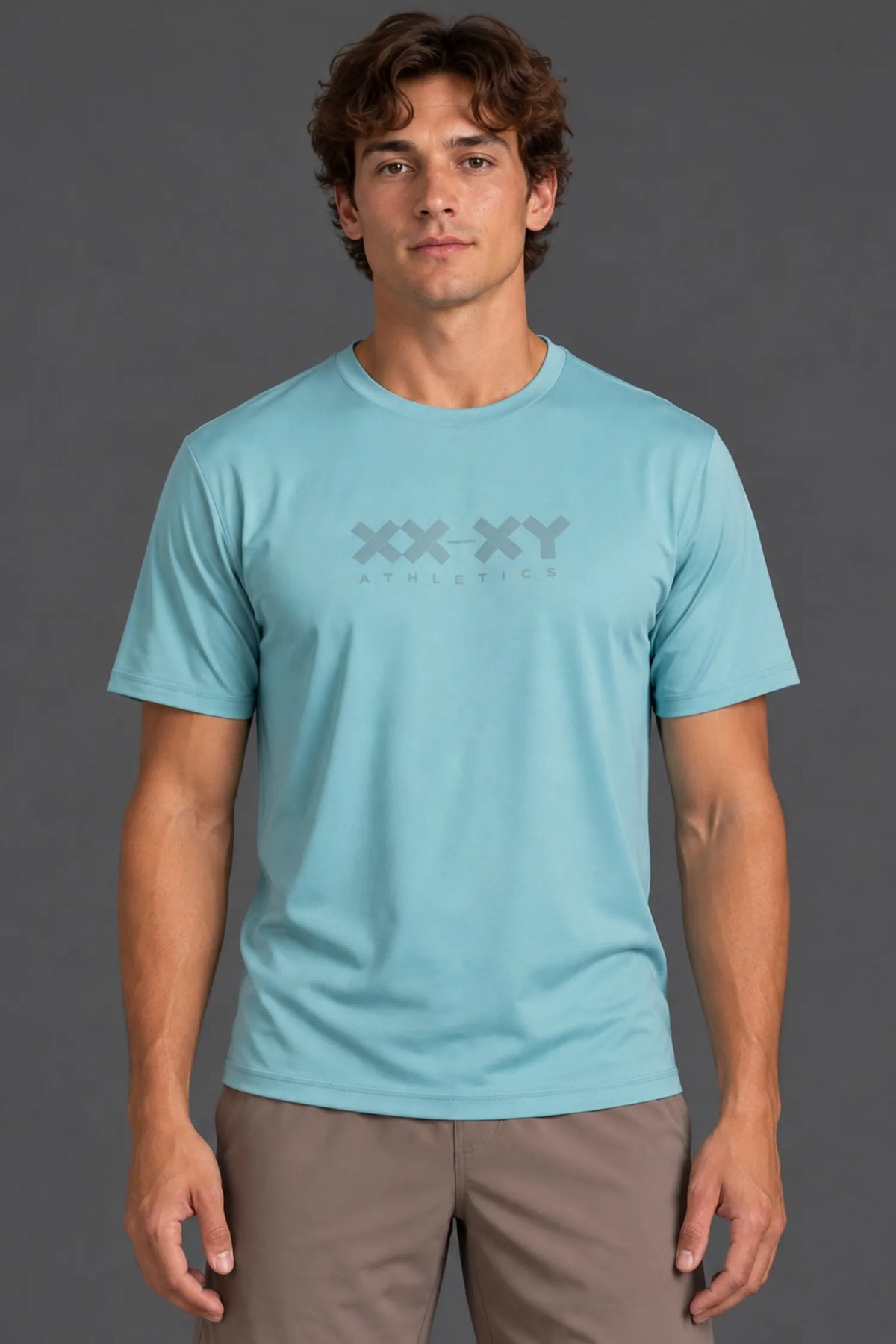 A man wearing a light blue crewneck athletic t-shirt with a gray 'XX-XY ATHLETICS' logo on the chest, paired with brown pants, against a dark gray studio background.