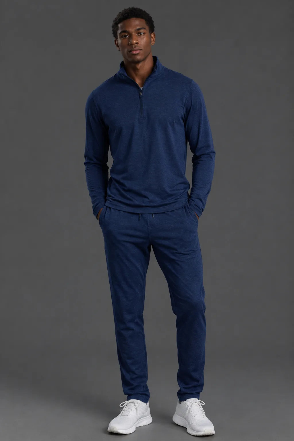 A man modeling a matching navy blue quarter-zip pullover and jogger pants set, paired with white sneakers, against a solid dark gray studio background.